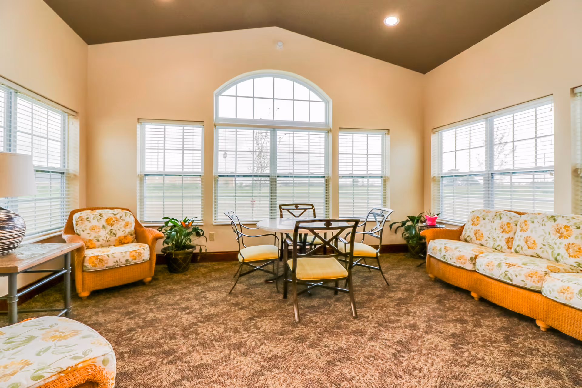 Bright communal sitting room with floral-upholstered wicker sofa and armchairs arranged around a round table in front of large multi-pane windows.