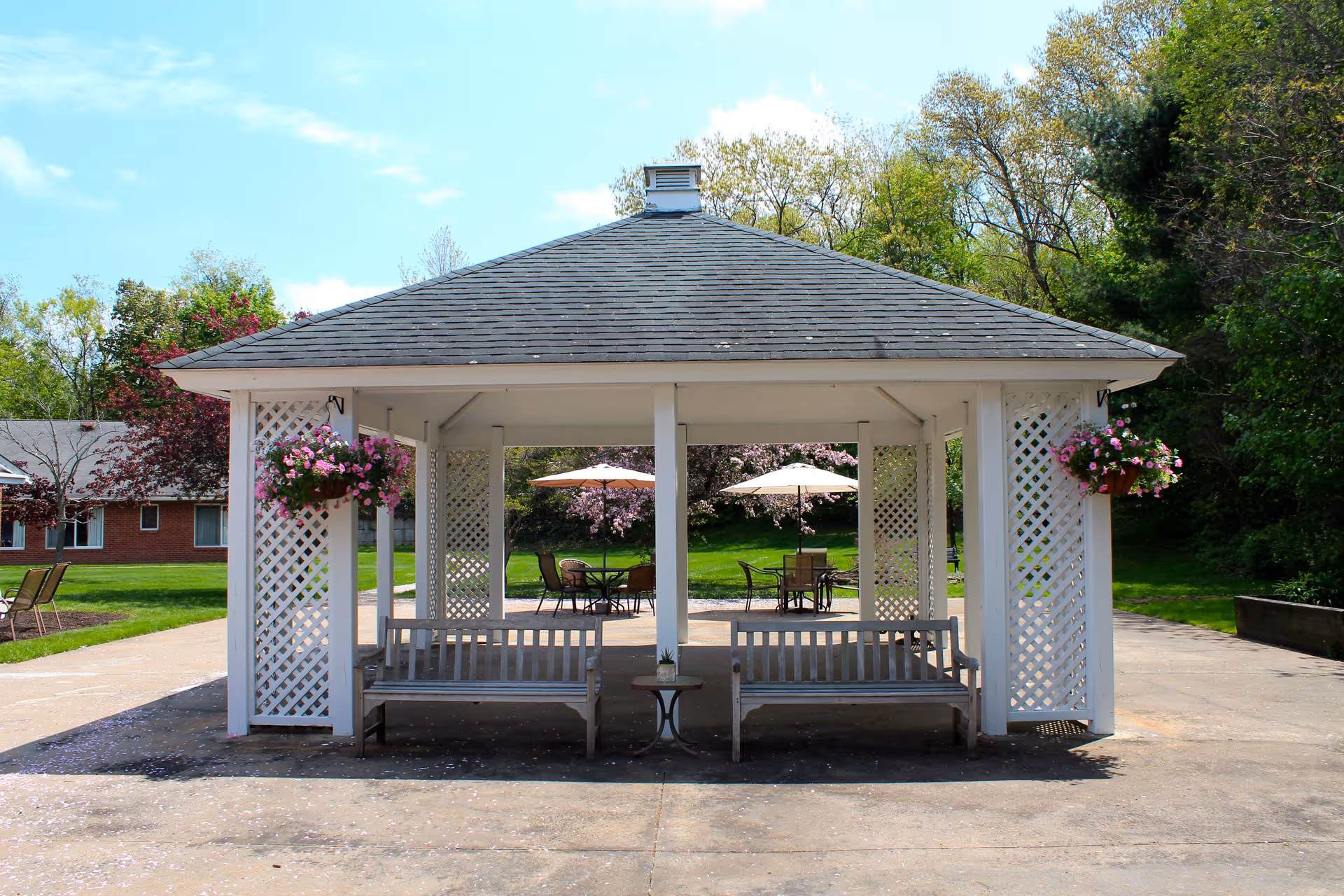 A white gazebo with a shingled roof and lattice sides, featuring two wooden benches and a small table inside. Hanging flower baskets with pink flowers are attached to the gazebo. In the background, there are patio tables with umbrellas, green grass, trees, and a brick building.