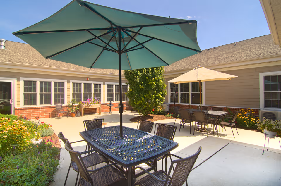 Outdoor courtyard area at Parkside Manor Assisted Living and Memory Care featuring patio tables with umbrellas, surrounded by a garden with flowers and shrubs, and the building with multiple windows in the background under a clear blue sky.