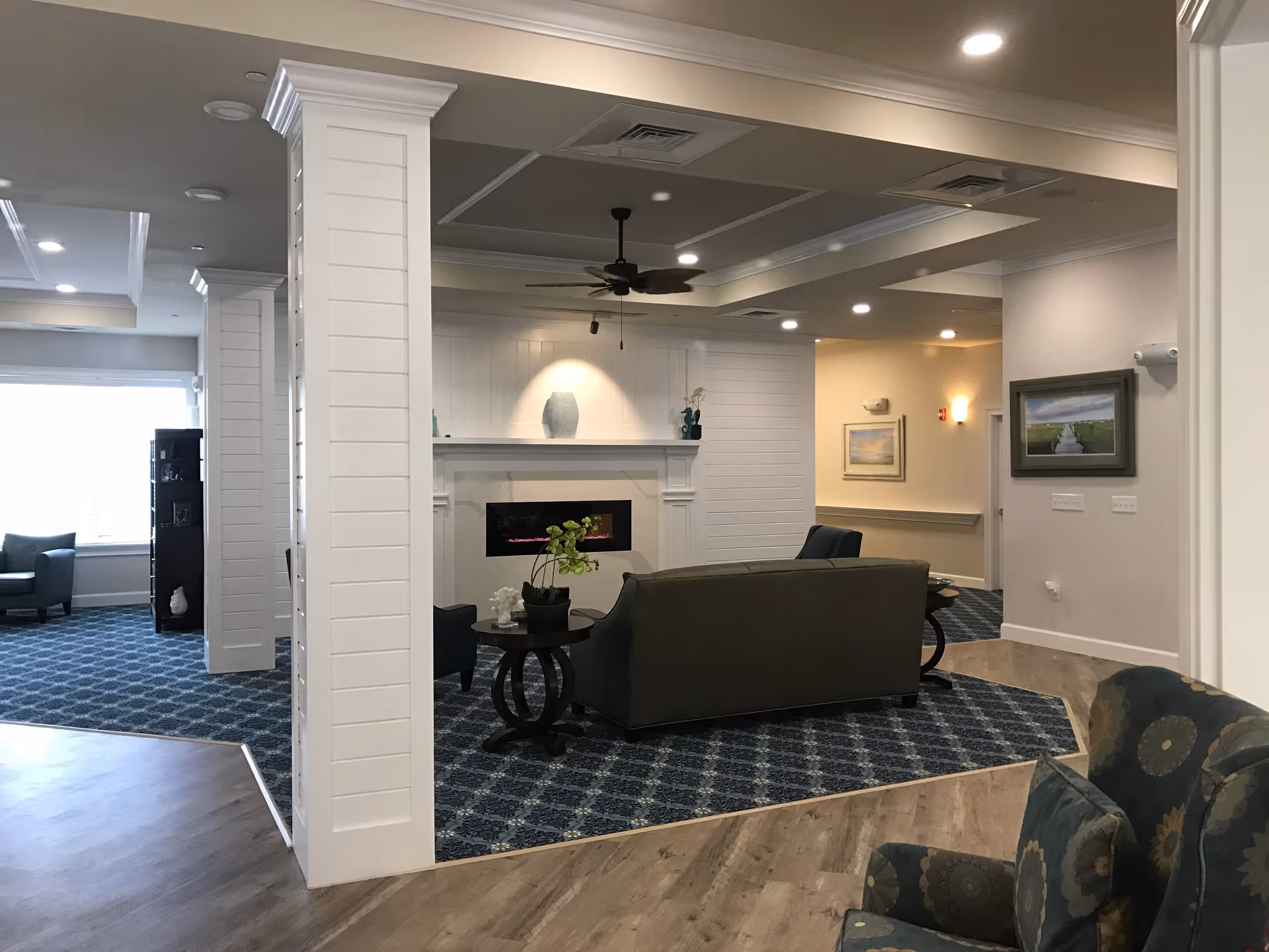 Interior view of a senior living facility lounge area with a patterned blue carpet, a gray sofa, armchairs, a small round table with a plant, a modern fireplace, white paneled columns, ceiling fan, and framed artwork on the walls.