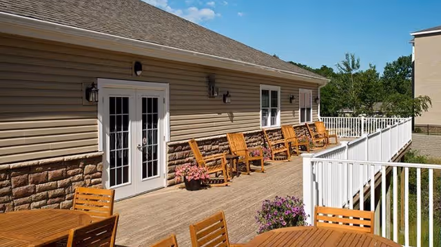 Outdoor patio area at Glenmont Abbey Village with wooden chairs and tables on a wooden deck attached to a beige building with white-framed glass doors and windows, under a clear blue sky.