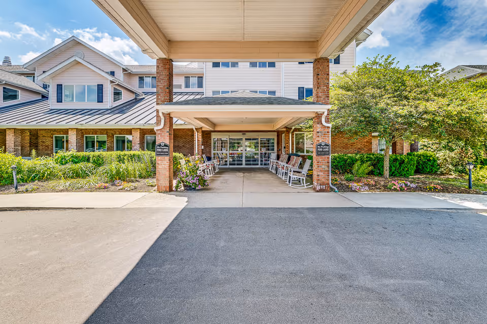 Entrance of Solstice Senior Living at Bangor showing a covered driveway with brick pillars, rocking chairs lined up on both sides, landscaped greenery and flowers, and a multi-story building in the background under a blue sky with clouds.