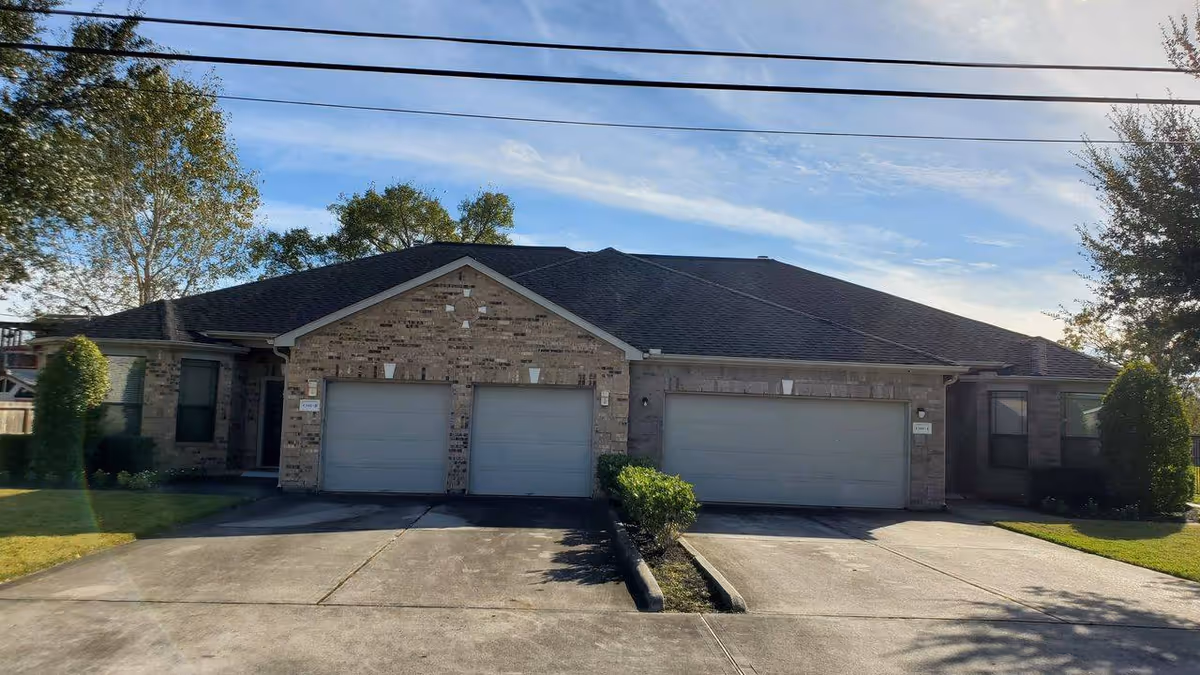 Single-story brick assisted living building front with three garage doors, driveways, and small landscaped lawns under a blue sky.