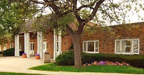Exterior view of a single-story brick building with white-framed windows and a covered entrance supported by white columns. There is a tree and landscaped bushes and flowers in front of the building.