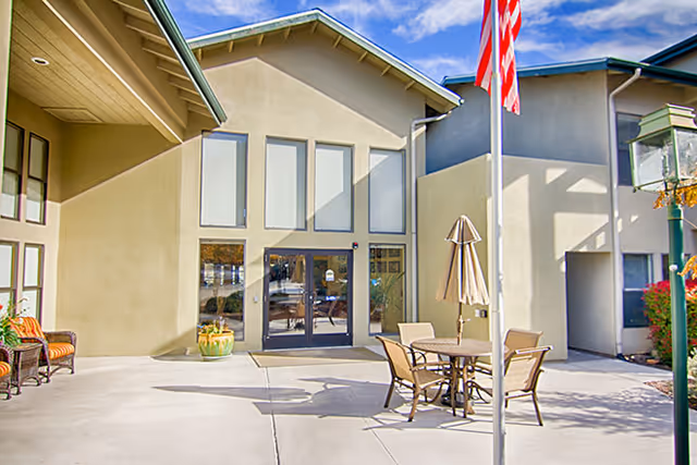 Outdoor patio area of a senior living facility with a round table and four chairs, an umbrella, a flagpole with an American flag, and a building entrance with large windows and double glass doors.
