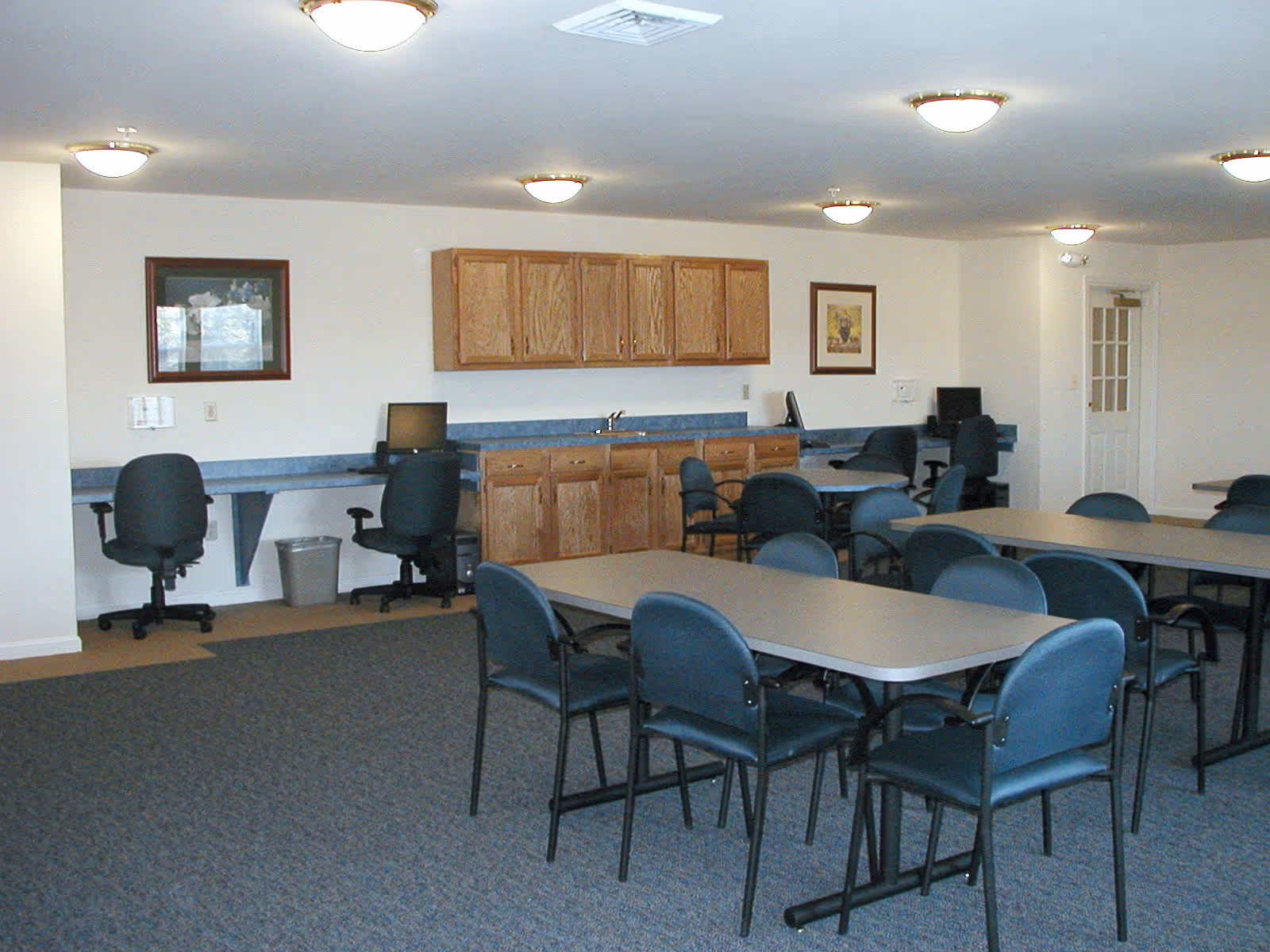 A spacious room with multiple tables and chairs arranged for group seating. Along one wall, there are wooden cabinets above and below a countertop with a sink. There are also several computer workstations with office chairs. The room has carpeted flooring and ceiling lights.