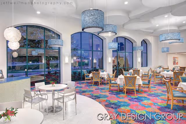 A brightly lit dining room with large arched windows, colorful patterned carpet, and multiple tables covered with white tablecloths. The room features modern hanging light fixtures and a mix of wooden and metal chairs. Outside the windows, some greenery is visible.
