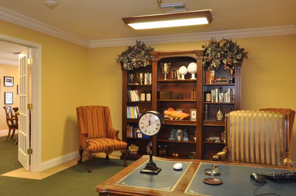 A cozy office-like sitting area with a wooden bookcase, two upholstered chairs, and a desk topped with a clock.