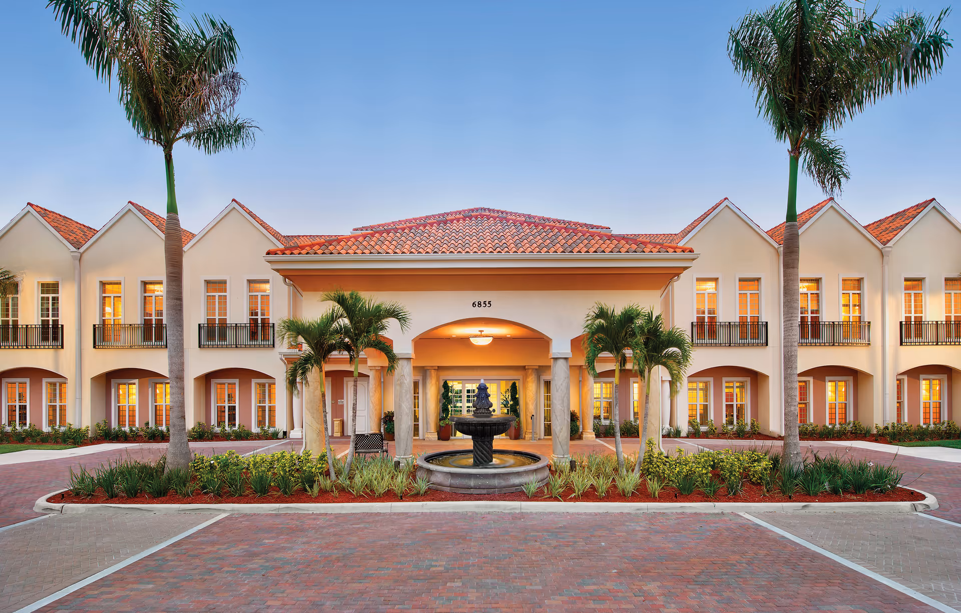 Mediterranean-style two-story senior living facility entrance with a central fountain, palm trees, and a tiled roof.