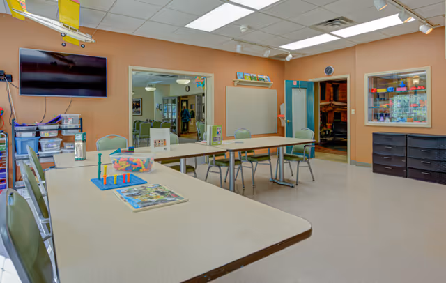 A bright activity room with beige walls and a tiled ceiling. Several tables and green chairs are arranged in the room, with board games and colorful toys on the tables. A large flat-screen TV is mounted on the wall to the left, above storage bins filled with supplies. There is a window on the right wall displaying various colorful items, and two doorways lead to adjacent rooms with more chairs and tables visible.
