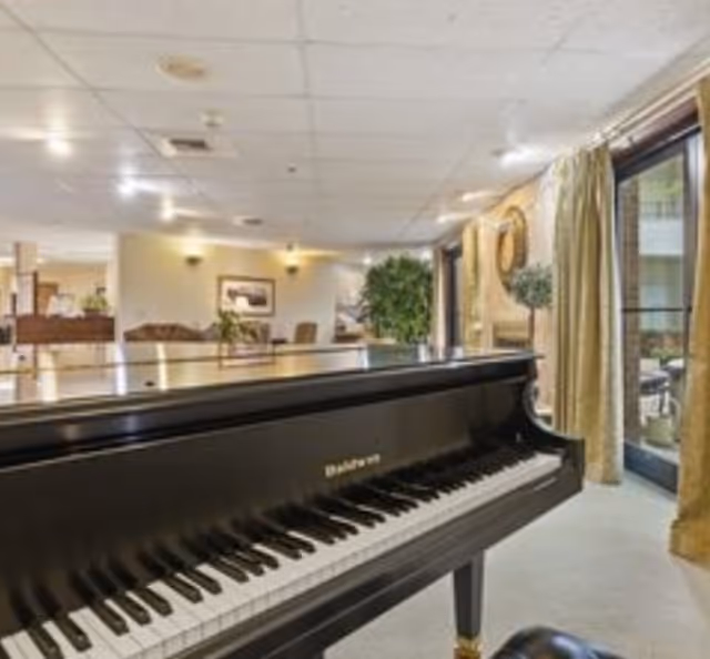 Interior view of a senior living facility common area featuring a black Baldwin grand piano in the foreground, with seating areas, plants, framed artwork on the walls, and large windows with curtains allowing natural light to enter.