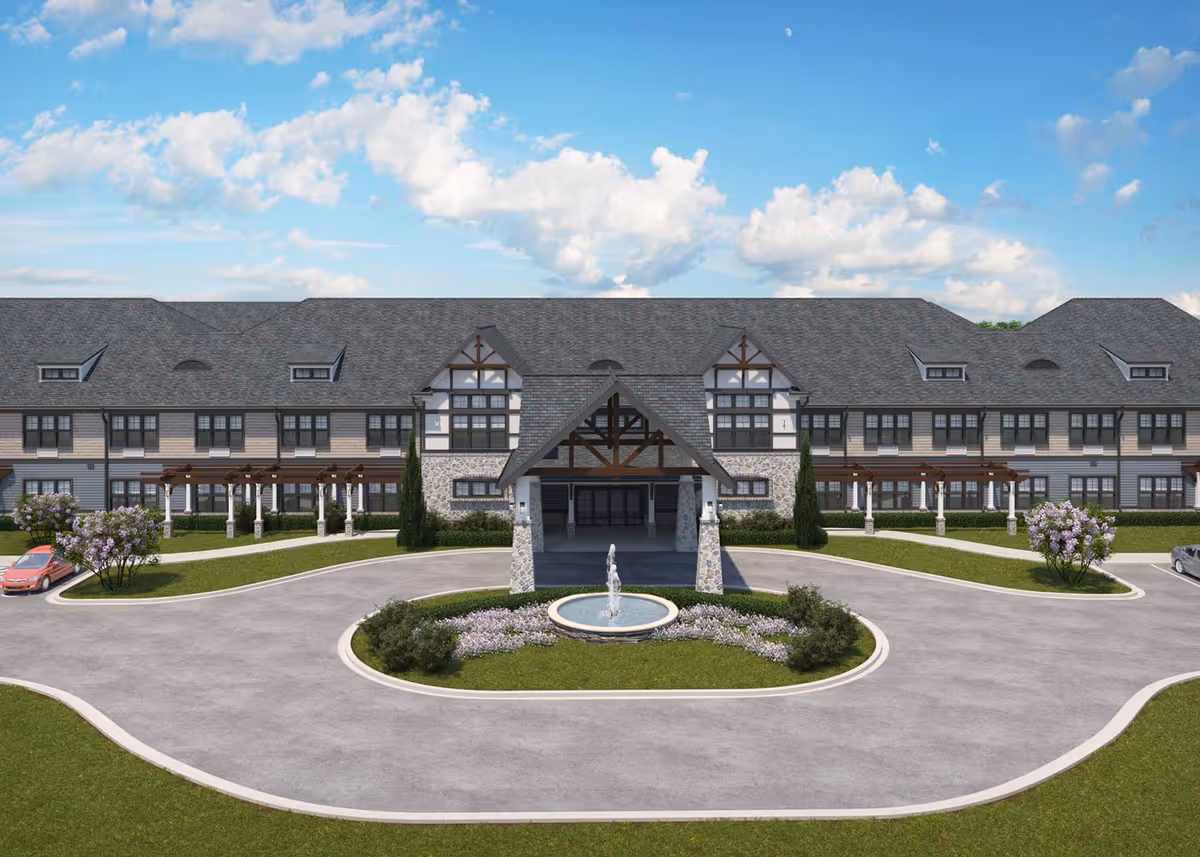 Front exterior view of a large senior living facility building with a covered entrance, stone pillars, a circular driveway with a fountain in the center, and landscaped greenery including bushes and flowering trees under a partly cloudy blue sky.