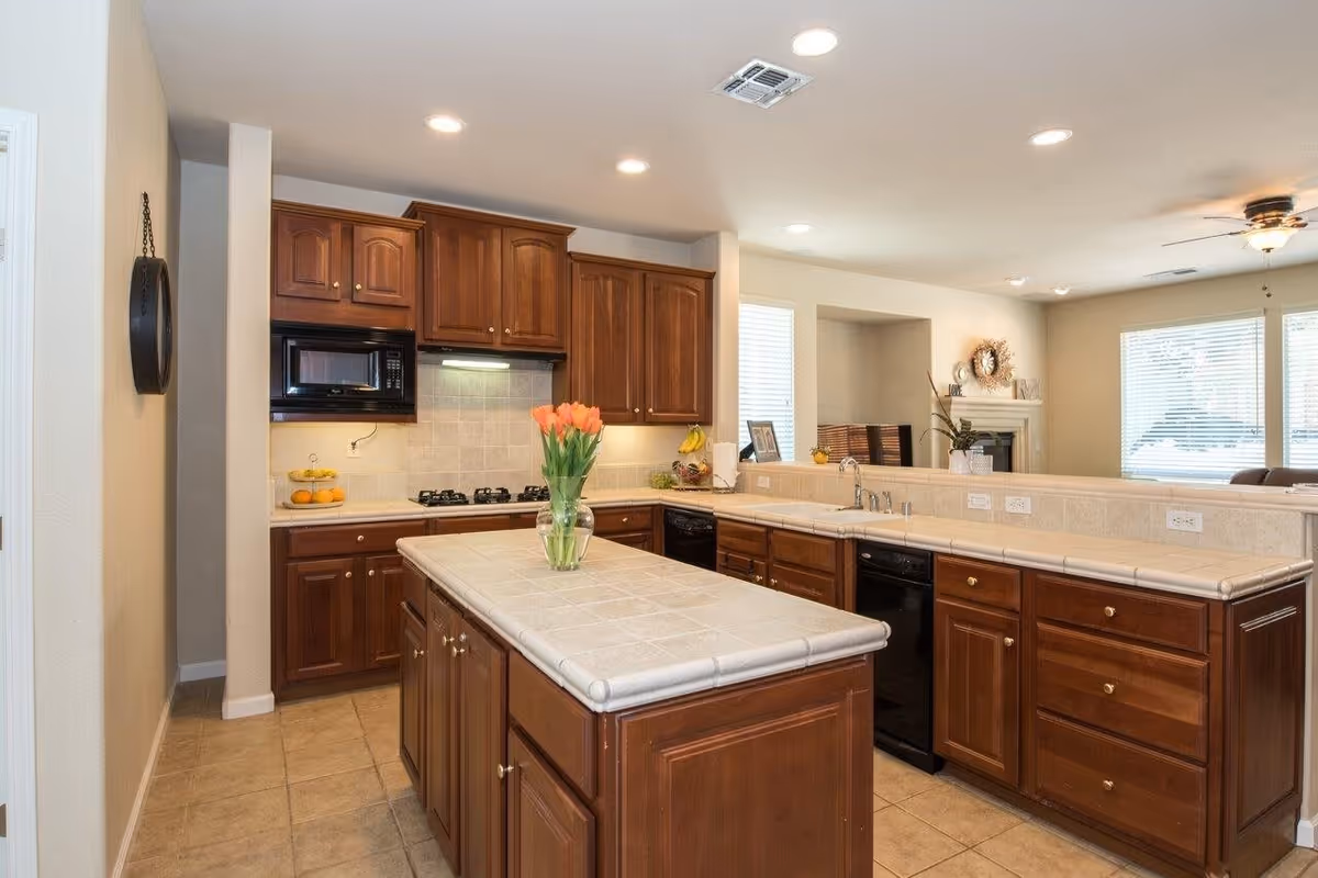 A spacious kitchen with wooden cabinets, a tiled countertop island with a vase of orange tulips, a built-in microwave, a gas stove, and a double sink. The kitchen opens into a living area with large windows and a ceiling fan.