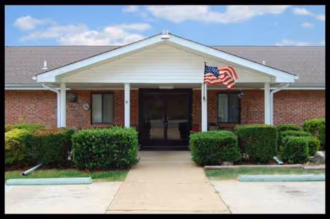 Front entrance of a brick senior living building with white columns, hedges, and an American flag.