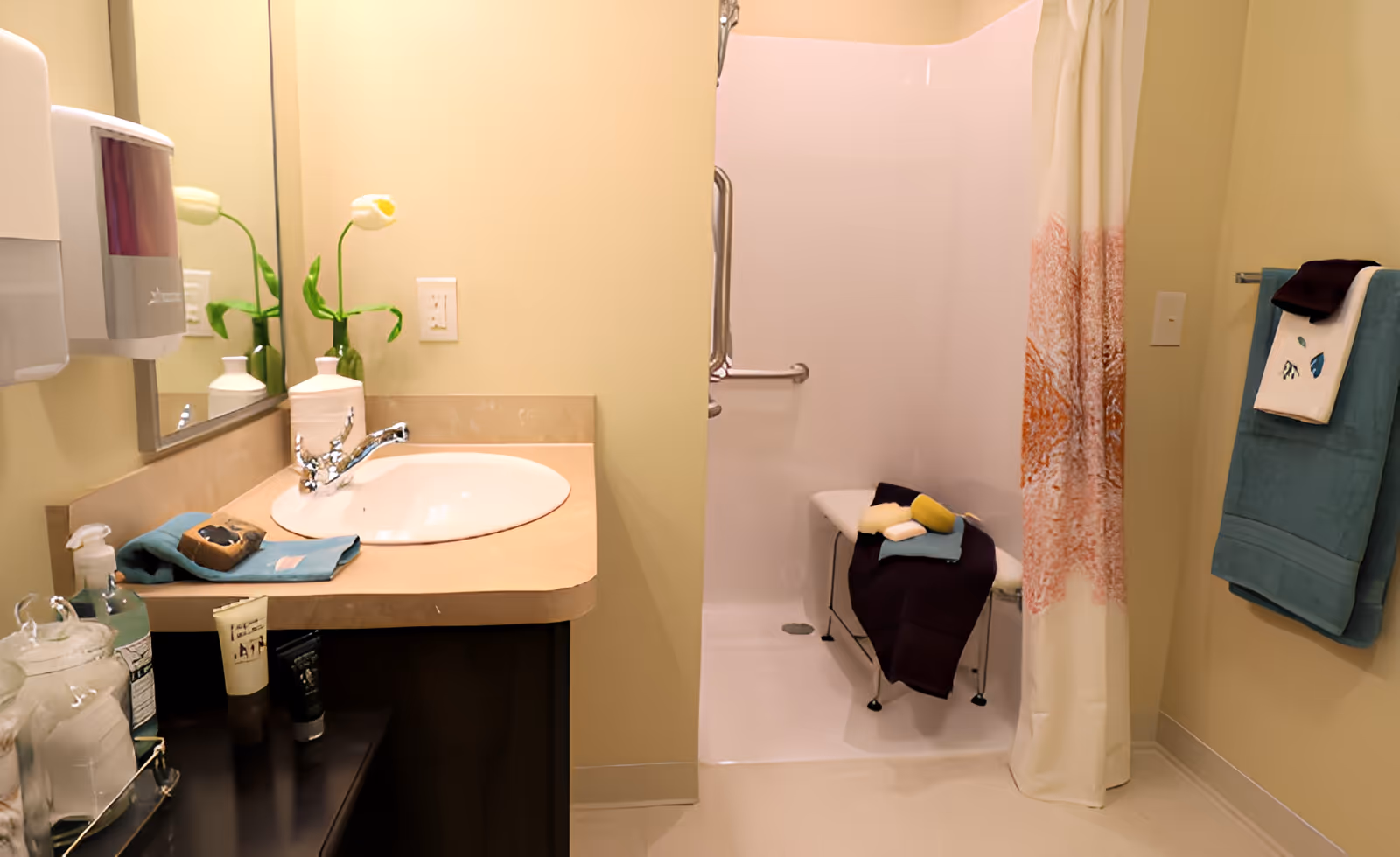 A clean and accessible bathroom with a beige countertop sink, a mirror, and a soap dispenser. There is a walk-in shower with a white curtain featuring an orange pattern, a shower bench with folded towels and sponges, and a towel rack with teal and white towels on the wall.