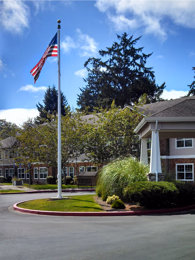 Front entrance of a senior living facility with an American flag on a flagpole, landscaped circular driveway and covered portico.