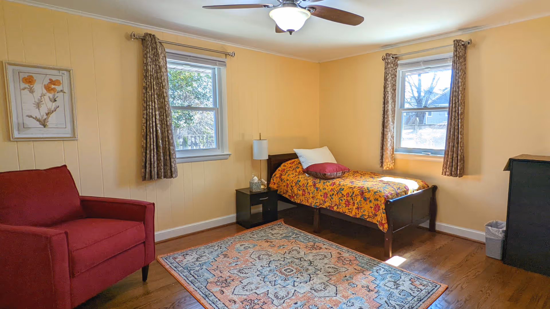 A cozy bedroom with yellow walls featuring a single bed with a colorful floral bedspread and pillows, a red armchair, a patterned area rug on wooden floor, two windows with brown curtains, a small black nightstand with a lamp and tissue box, a black dresser, and a ceiling fan with light.