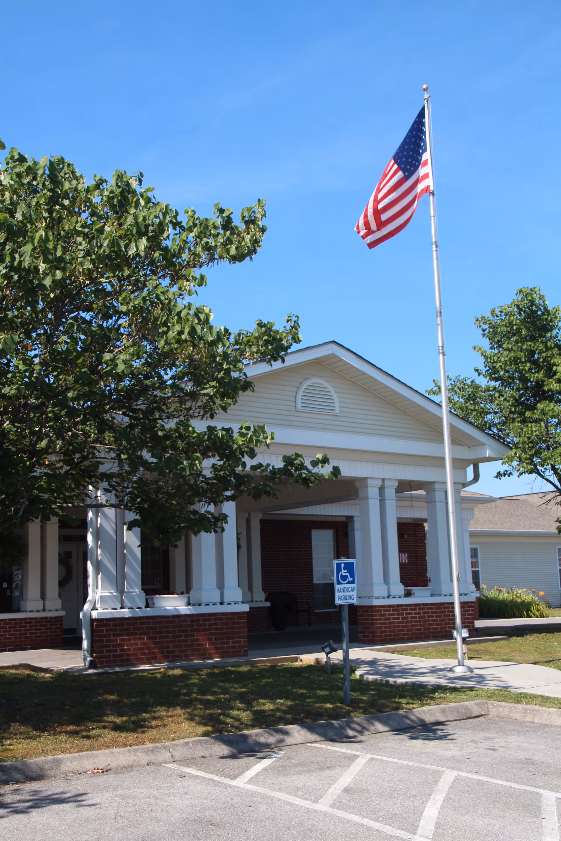 Entrance of Wellington Manor Assisted Living facility showing a covered porch with white columns and brick base, an American flag on a flagpole, a handicap parking sign, and trees under a clear blue sky.
