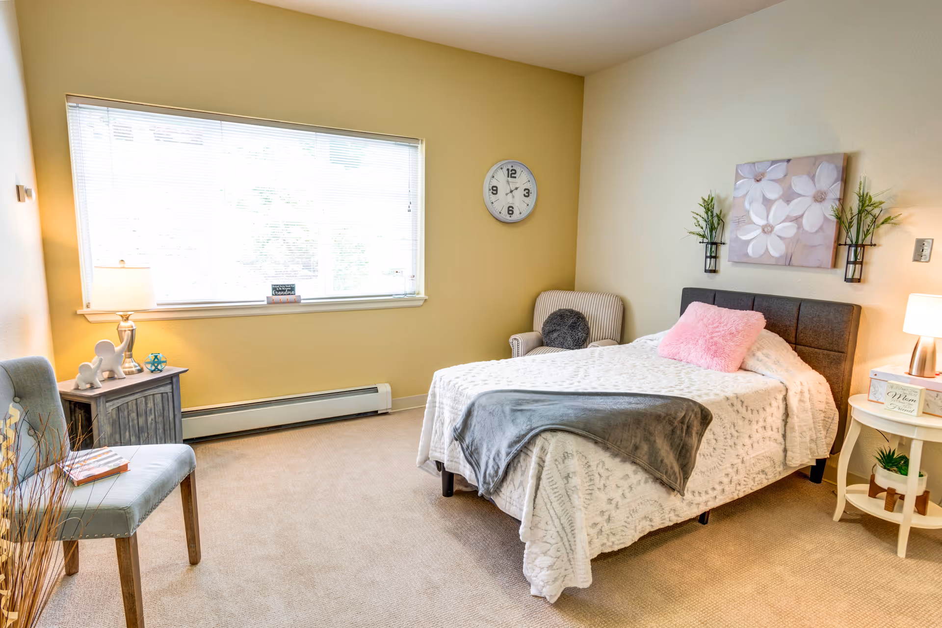 Bright, cozy bedroom with a made bed, two chairs, nightstands with lamps, wall decor, and a large window.