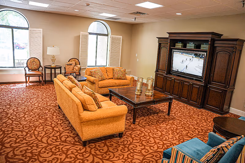 A cozy senior living common area with two orange sofas, a wooden coffee table with decorative candles, a large wooden entertainment center with a TV showing a hockey game, two chairs near windows with white shutters, and a patterned red carpet.