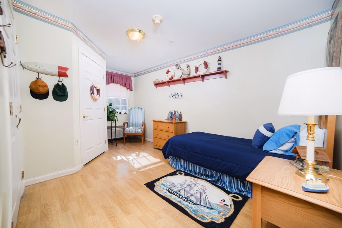 A small bedroom with a single bed covered in a navy blue comforter and blue pillows. There is a wooden nightstand with a lamp and a small model ship on it. A wooden dresser is against the wall with nautical-themed decorations on top and a red shelf above holding life preservers, an anchor, and a lighthouse figurine. A striped armchair is near a window with a purple valance. The floor is light wood with a nautical-themed rug featuring a sailing ship. Hats hang on hooks on the wall near a closed white door.