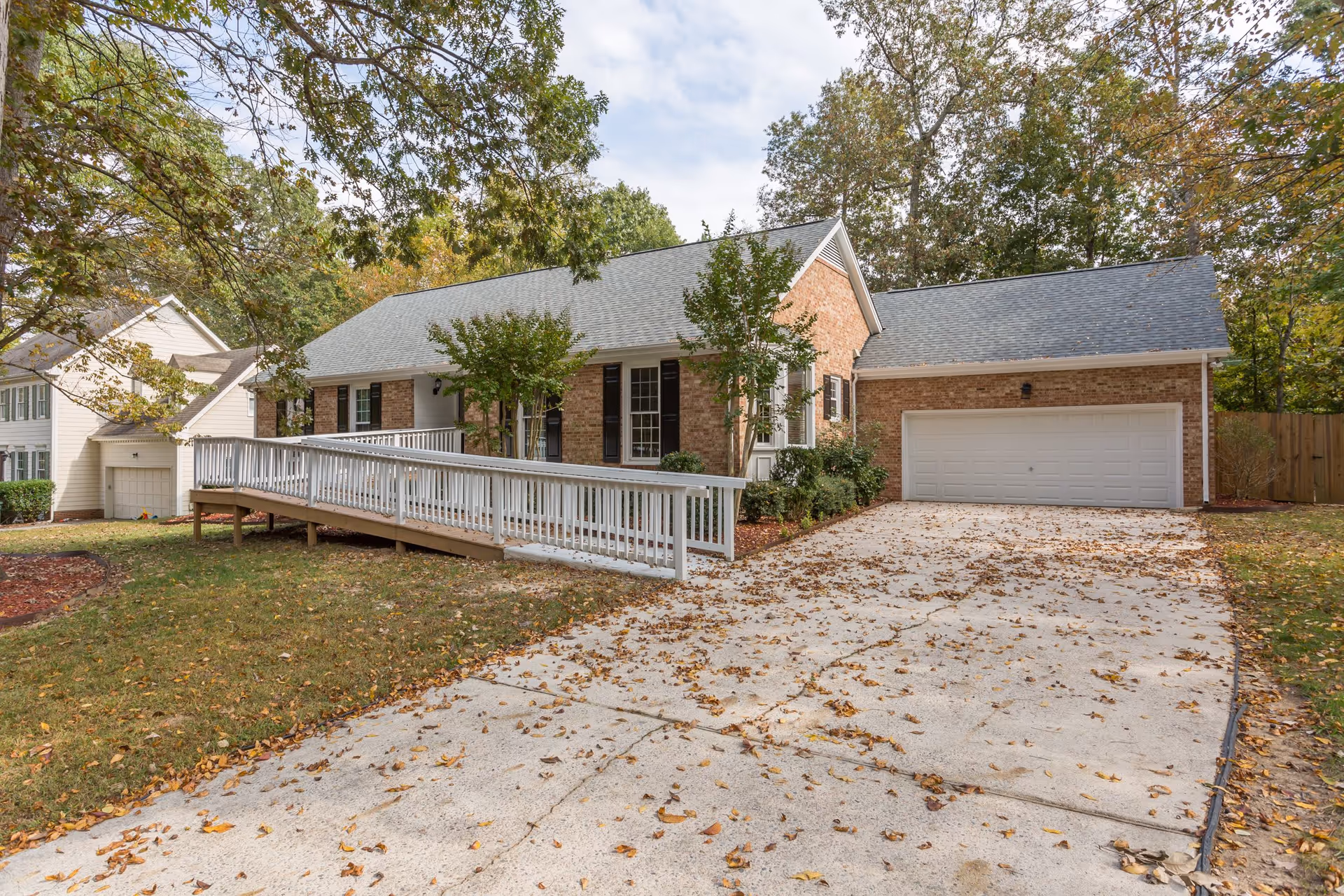Exterior view of a single-story brick assisted living facility with a gray shingled roof, white garage door, and a wheelchair-accessible ramp leading to the front entrance. The driveway and lawn are covered with fallen autumn leaves, and trees surround the property.