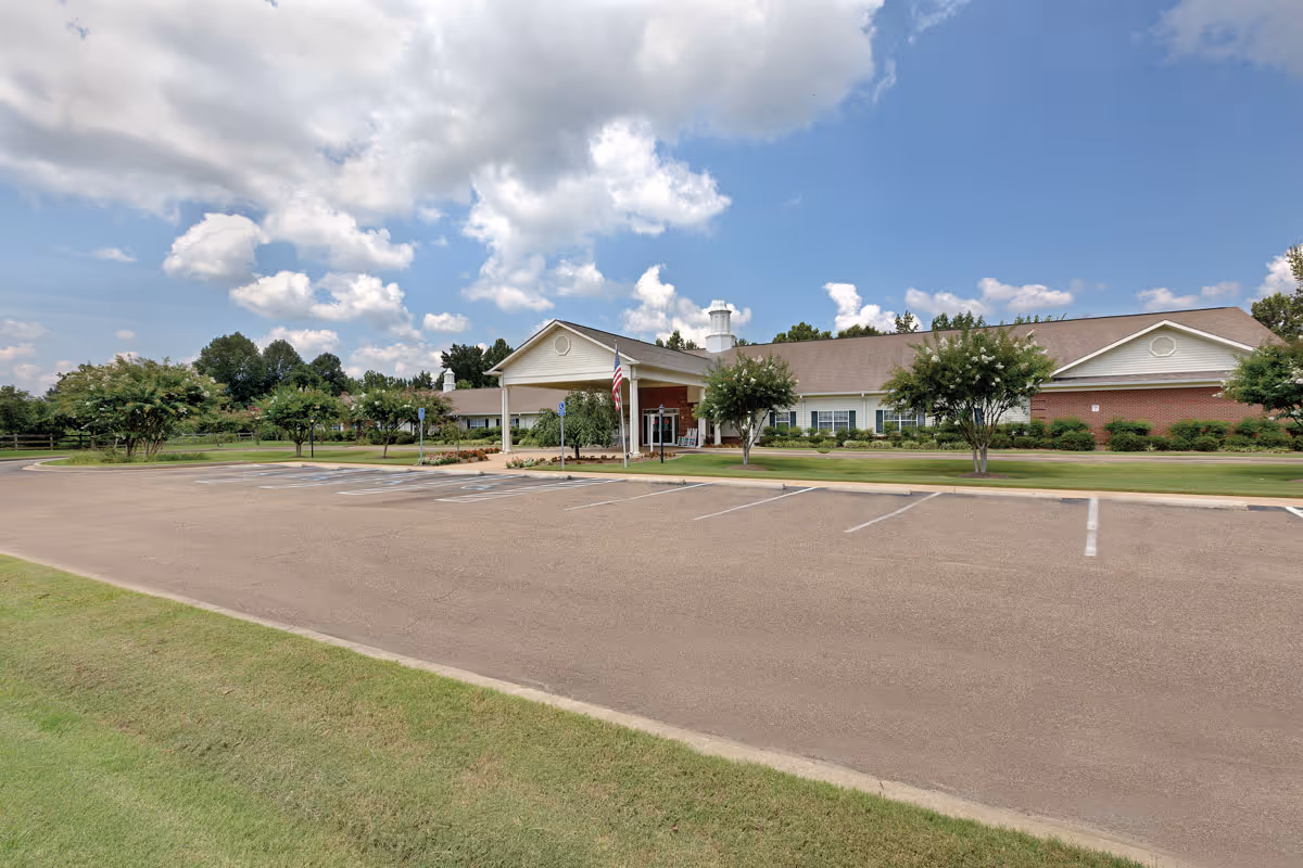 Exterior view of The Pinnacle of Oxford senior living facility showing a single-story building with a covered entrance, an American flag, several trees, and an empty parking lot under a partly cloudy sky.