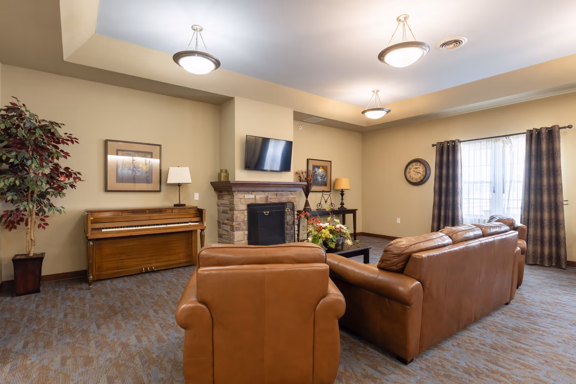 Cozy communal living room with leather sofas facing a fireplace and wall-mounted TV, a piano against the wall, and a large curtained window.