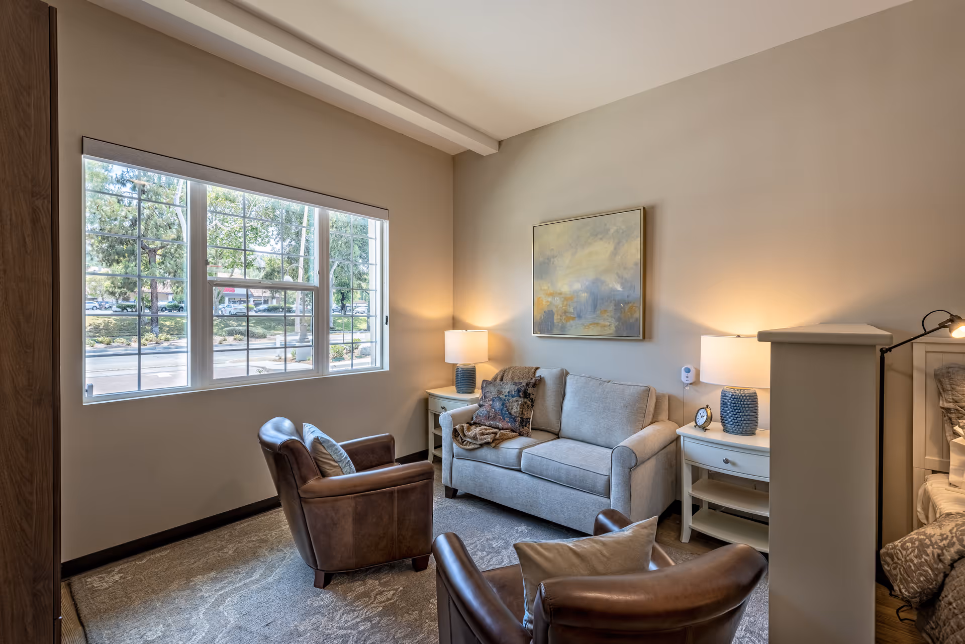 A cozy sitting area in a senior living facility with a beige loveseat, two brown leather armchairs, two side tables with lamps, a large window showing trees and a street outside, and a painting on the wall.