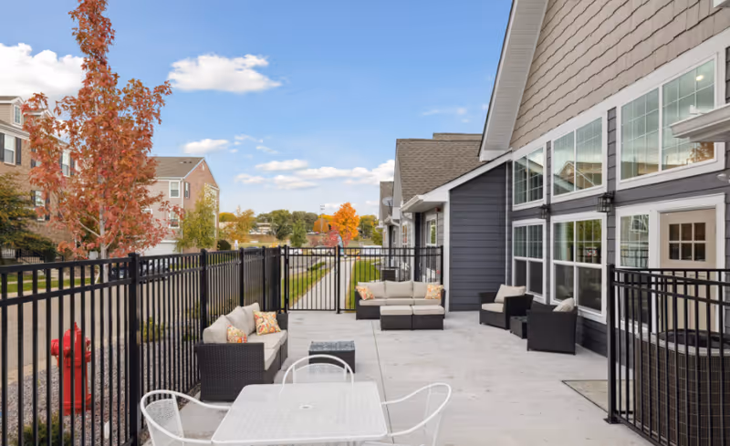 Outdoor patio with cushioned wicker seating, a metal table and chairs, and a black fence alongside the senior living building.