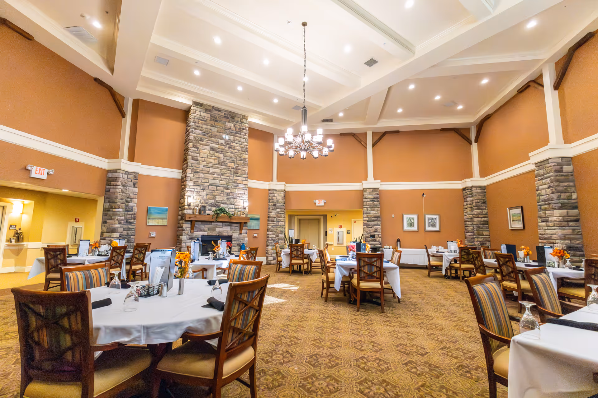 Spacious assisted living dining room with round tables set with white tablecloths, a stone fireplace, and a large chandelier under a high coffered ceiling.