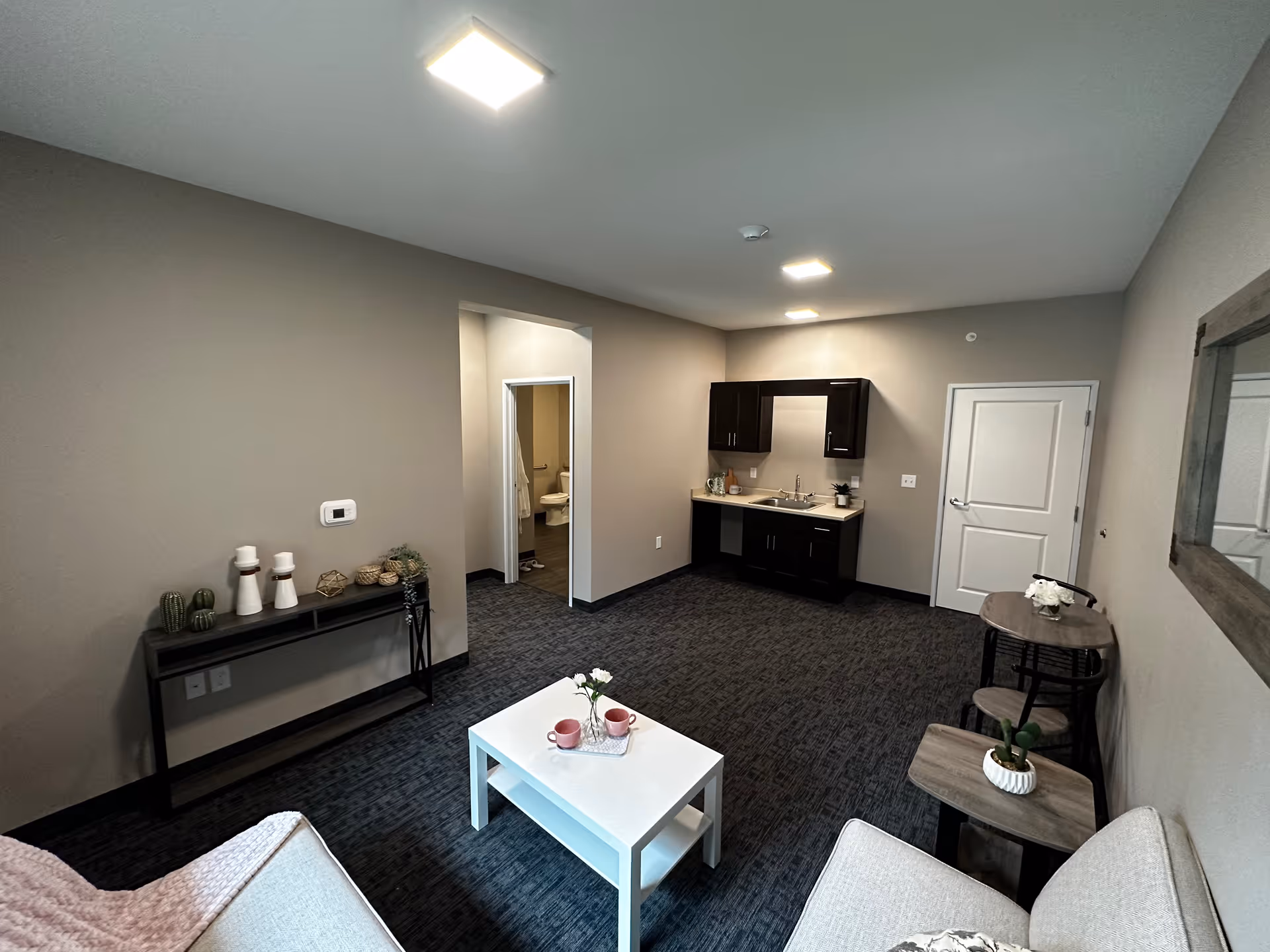 Interior view of a senior living facility room with a small kitchenette featuring dark cabinets and a sink. The room has a carpeted floor, a white coffee table with two pink cups and a small flower vase, a beige couch, a console table with decorative items, and a small round table with two chairs. A doorway leads to a bathroom with a visible toilet and bathrobe hanging on the wall.