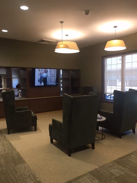 A cozy interior lounge area with three high-backed green upholstered chairs arranged around a small round table. A wall-mounted TV is visible on a wooden shelving unit, and two pendant lights hang from the ceiling. A large window allows natural light into the room.