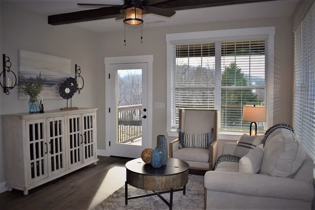 A cozy living room with a beige sofa and armchair, a round coffee table with decorative vases, a wooden cabinet with glass-paneled doors, a ceiling fan with lights, and large windows with white blinds letting in natural light. A door leads to an outdoor deck with a view of trees.