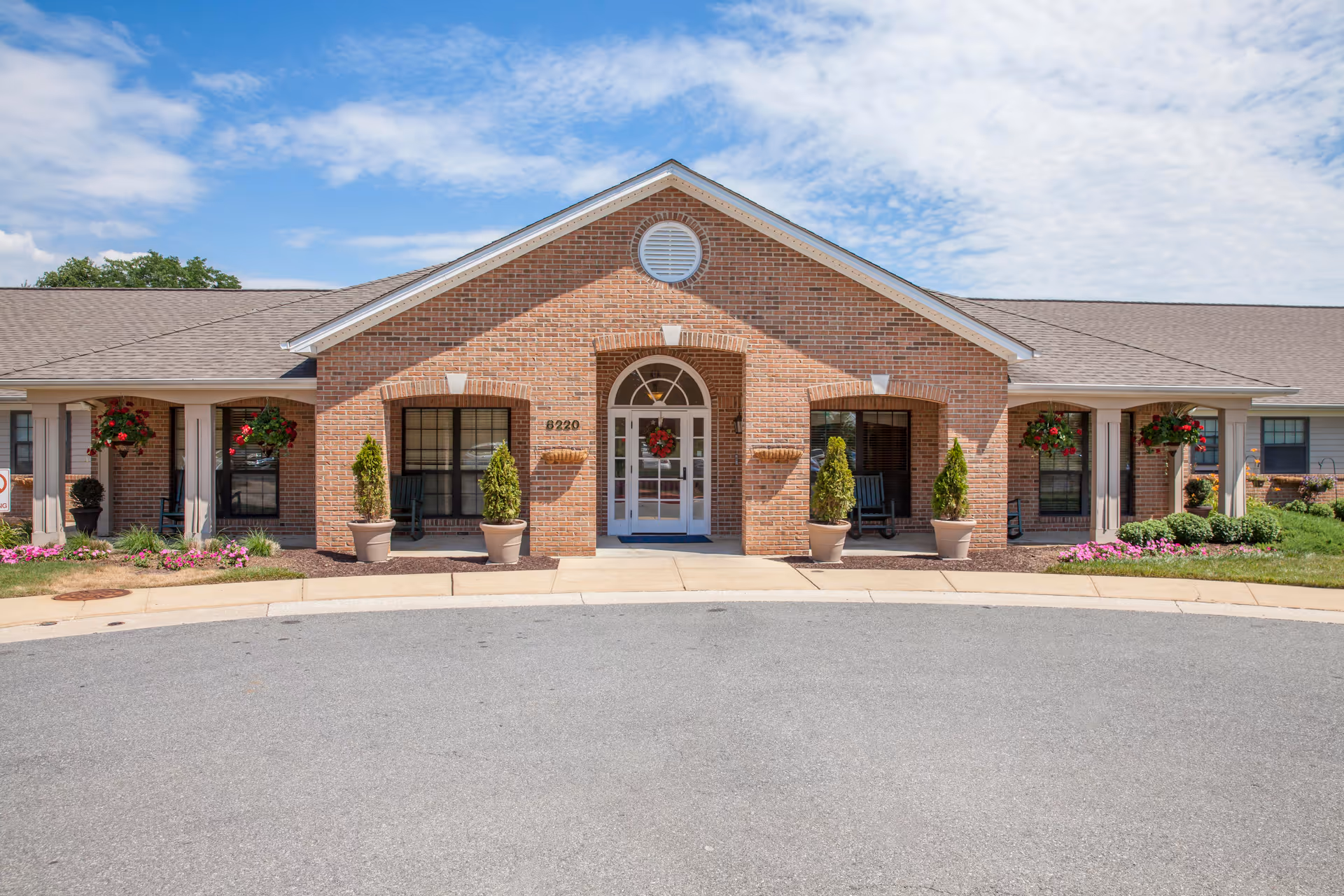 Front exterior view of a single-story brick building with a peaked roof, featuring a central entrance with glass doors decorated with a wreath. There are potted plants and hanging flower baskets on either side of the entrance, along with windows and rocking chairs. The building is surrounded by a paved driveway and landscaped with grass and flowers under a partly cloudy sky.