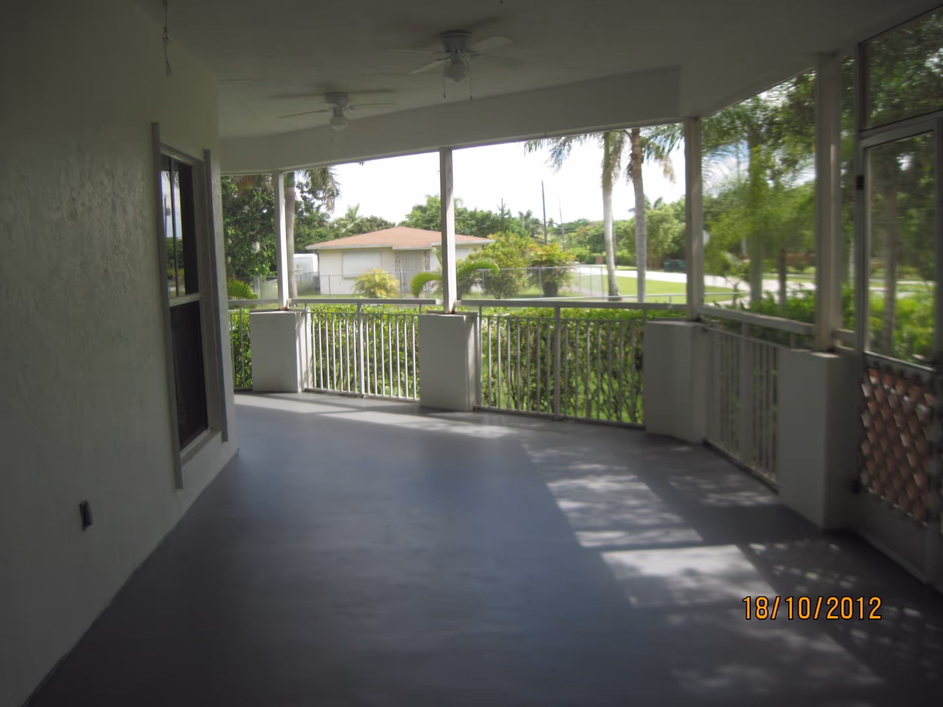 A spacious screened-in porch with a gray floor, white railings, and ceiling fans. The porch overlooks a green yard with palm trees and a neighboring house in the background.