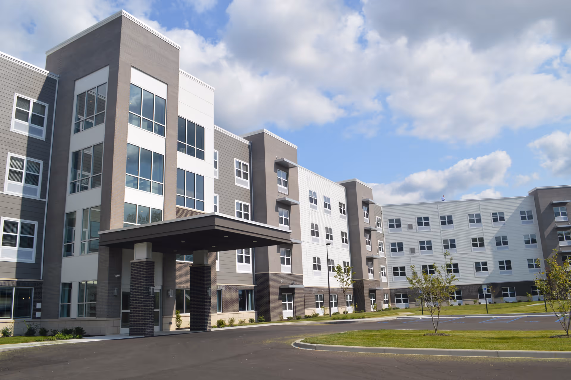 Exterior view of a modern multi-story senior living facility building with large windows, a covered entrance, and a parking area in front under a partly cloudy sky.