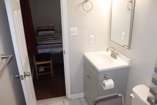 Small bathroom with a vanity and mirror, toilet with grab bar, and an open door showing a bed in the adjacent bedroom.