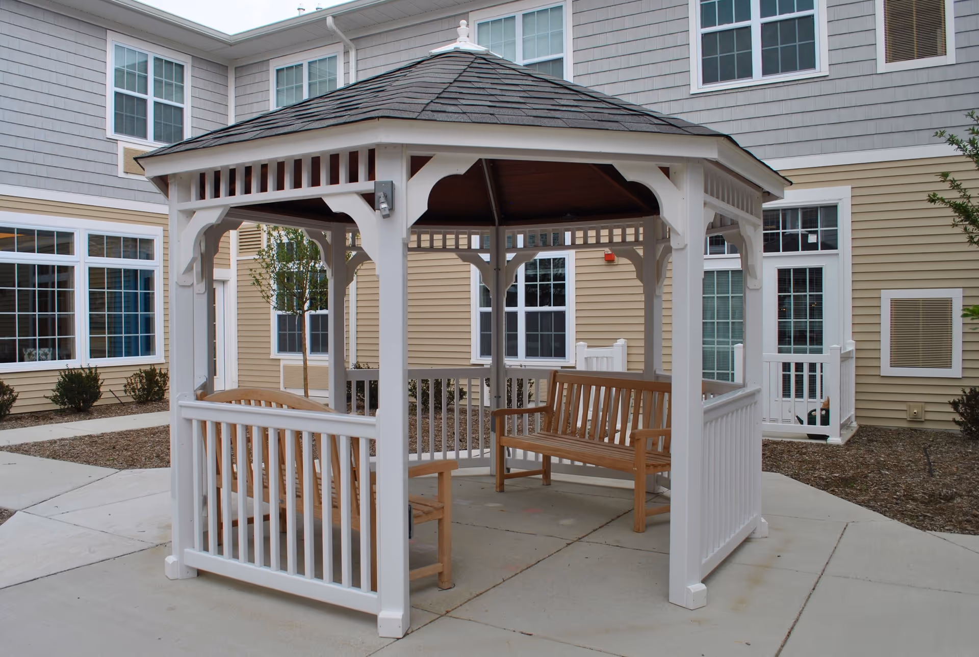 A white wooden gazebo with a shingled roof situated on a concrete patio outside a beige and gray building. Inside the gazebo are two wooden benches facing each other. The surrounding area includes windows, doors, small bushes, and a tree.