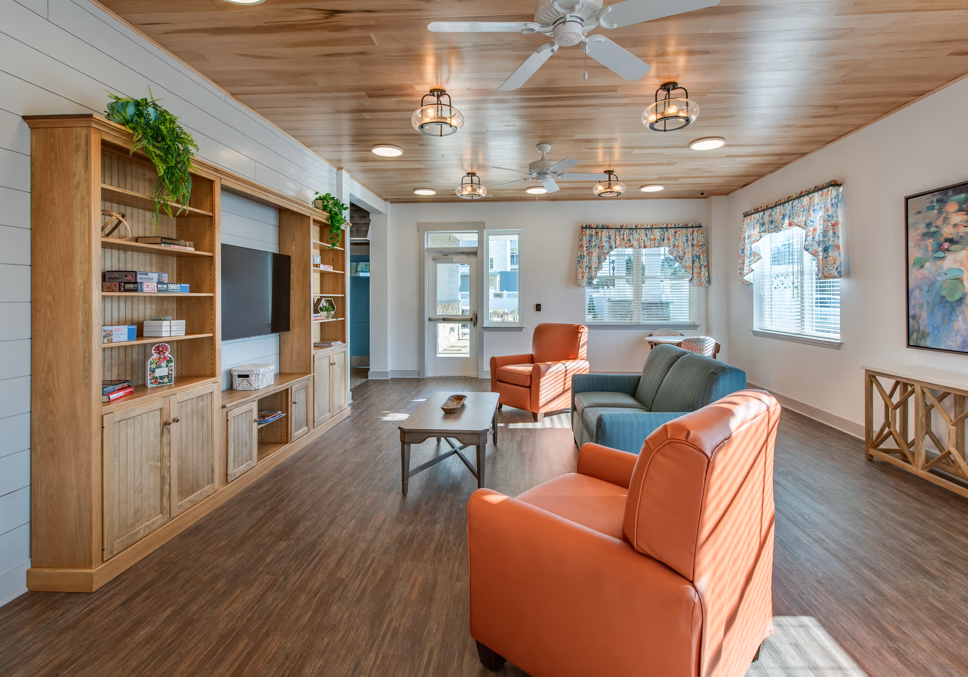 A bright and spacious living room with wooden flooring and ceiling, featuring a built-in wooden entertainment center with shelves and a flat-screen TV. The room has two orange armchairs, a green sofa, a wooden coffee table, and windows with floral valances letting in natural light. Ceiling fans and modern light fixtures are mounted on the ceiling.