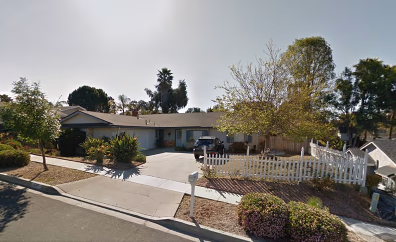 Single-story building front with a driveway, parked car, landscaping and a white picket fence.