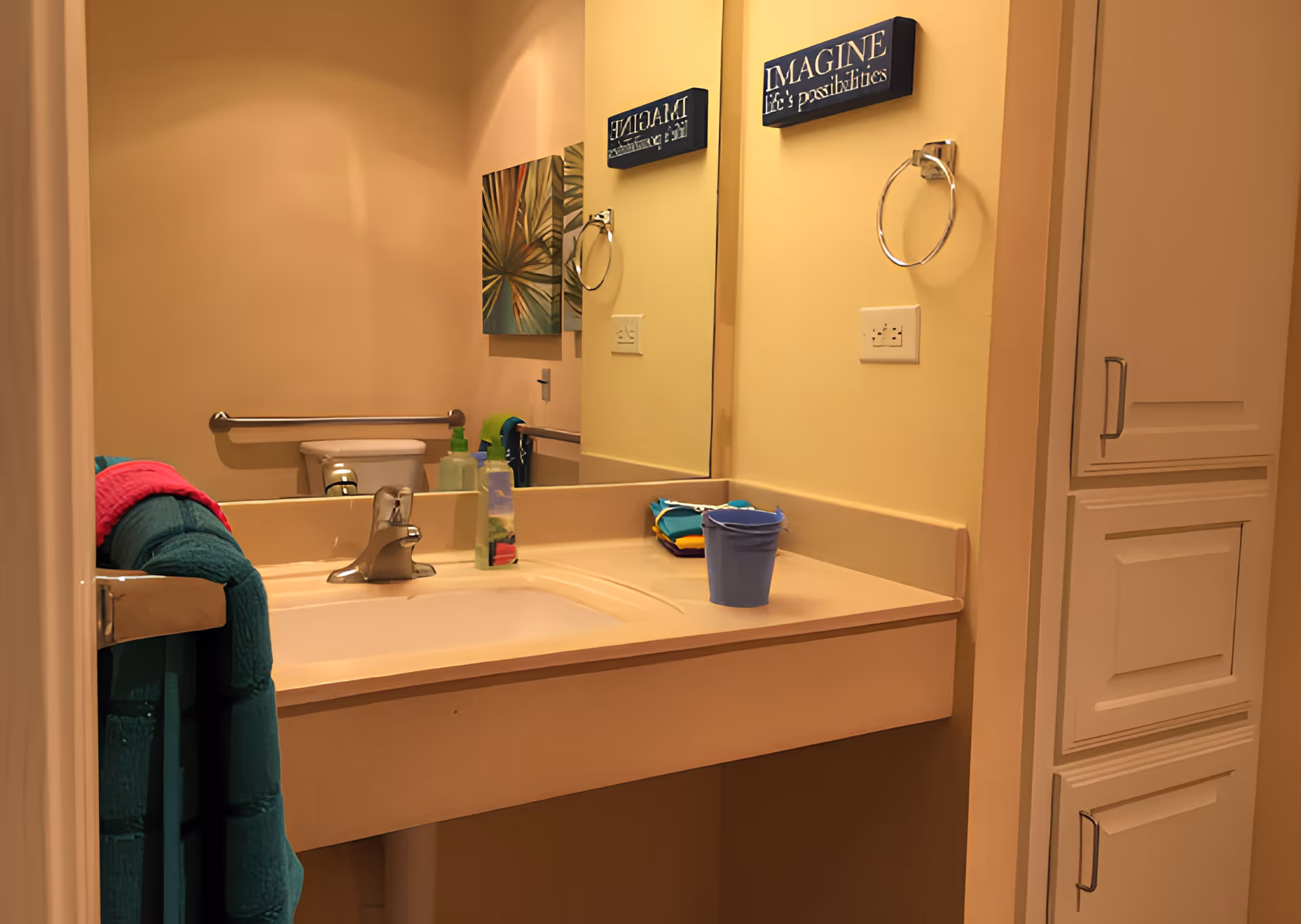 Accessible bathroom sink area with a mirror, faucet, towel, toiletries, grab bar and toilet visible in the background.