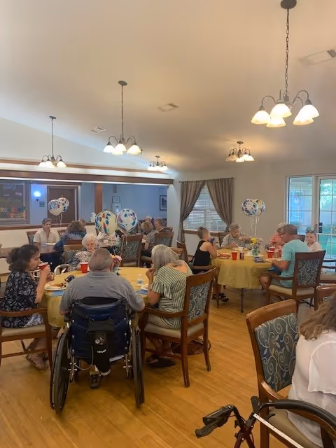 Seniors seated around tables with yellow tablecloths and balloons sharing a meal in a bright communal dining room.
