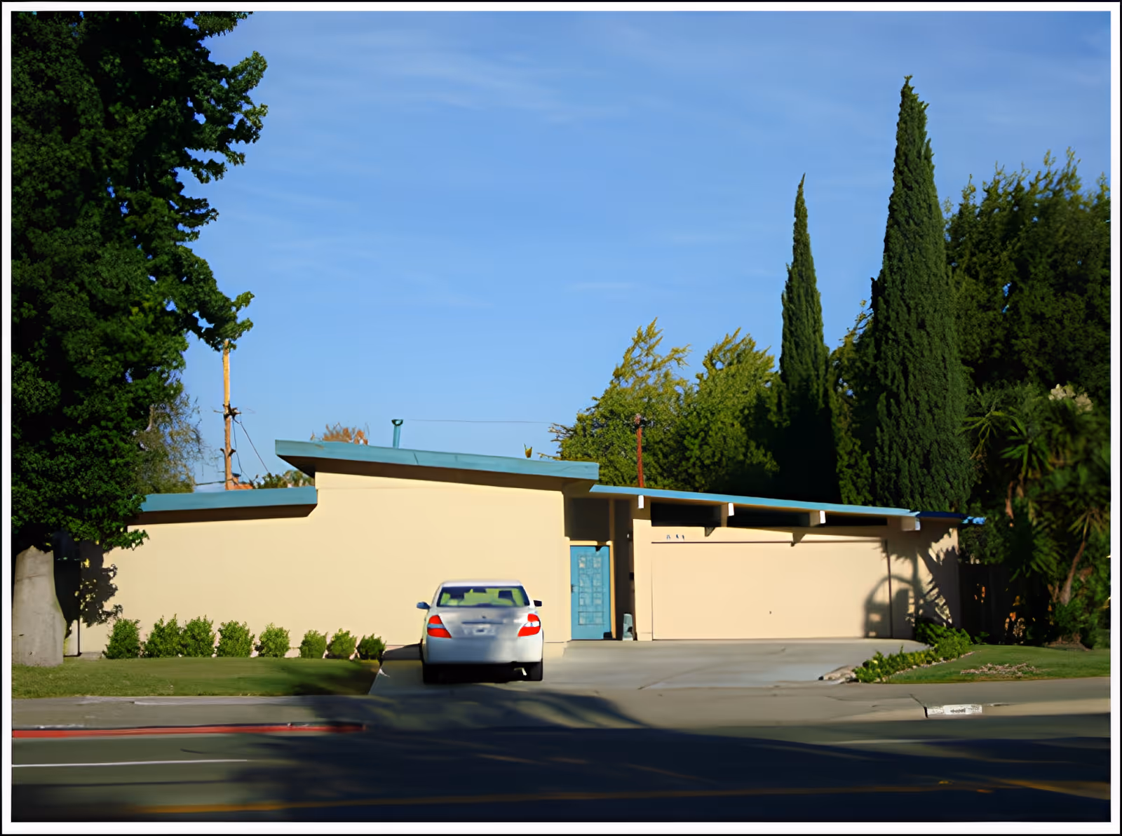 Front exterior view of a single-story residential building with a flat roof and a light beige facade. A silver car is parked in the driveway leading to a closed garage door. The entrance door is painted blue. The property is surrounded by green trees and shrubs under a clear blue sky.