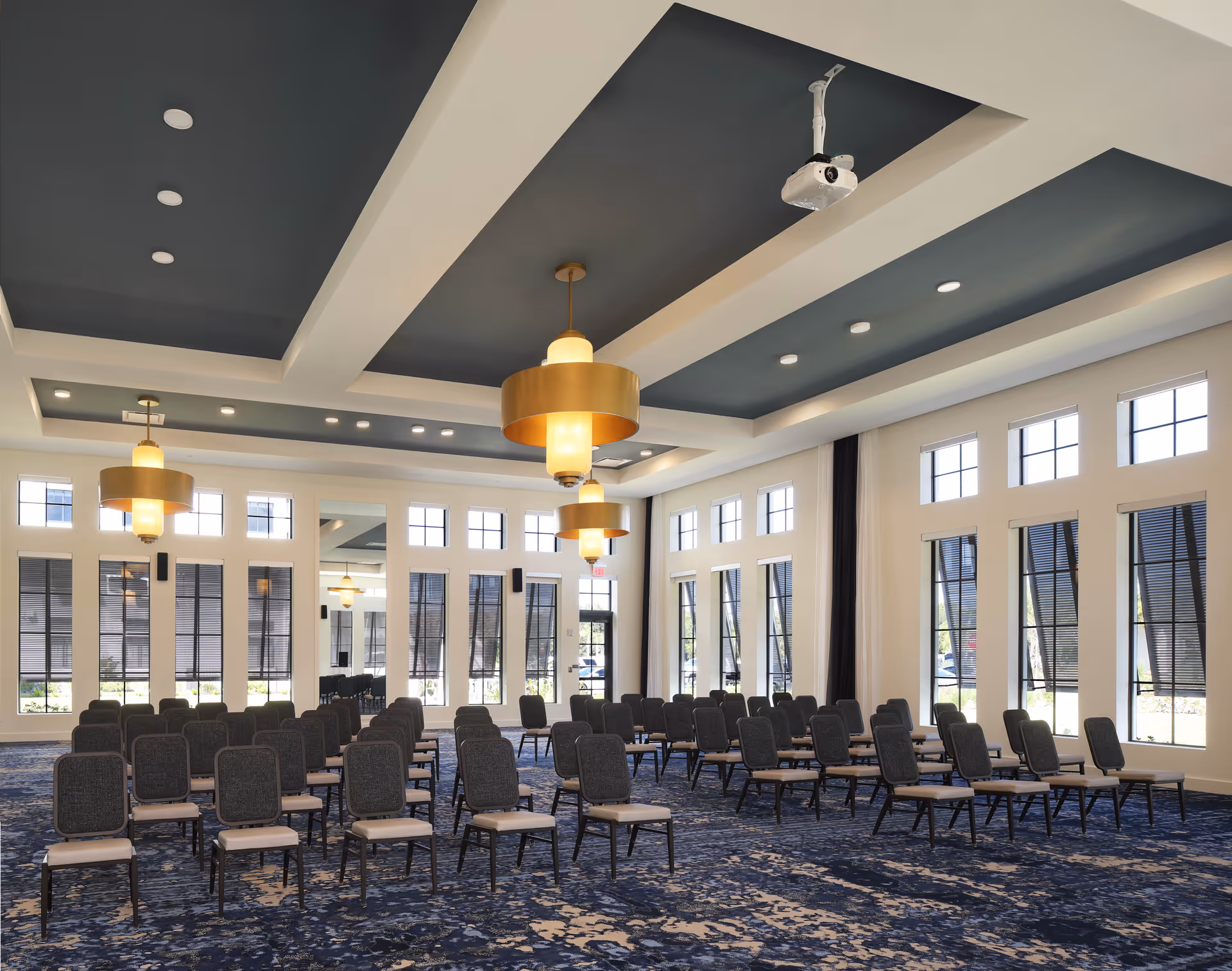 A spacious, well-lit conference or meeting room with rows of black chairs arranged facing forward. The room features large windows with black blinds, a patterned blue carpet, and modern ceiling lights with gold fixtures. A projector is mounted on the ceiling.