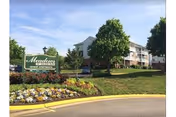 Entrance sign and landscaped front lawn with flowers, trees, and a multi-story senior apartment building with balconies under a blue sky.