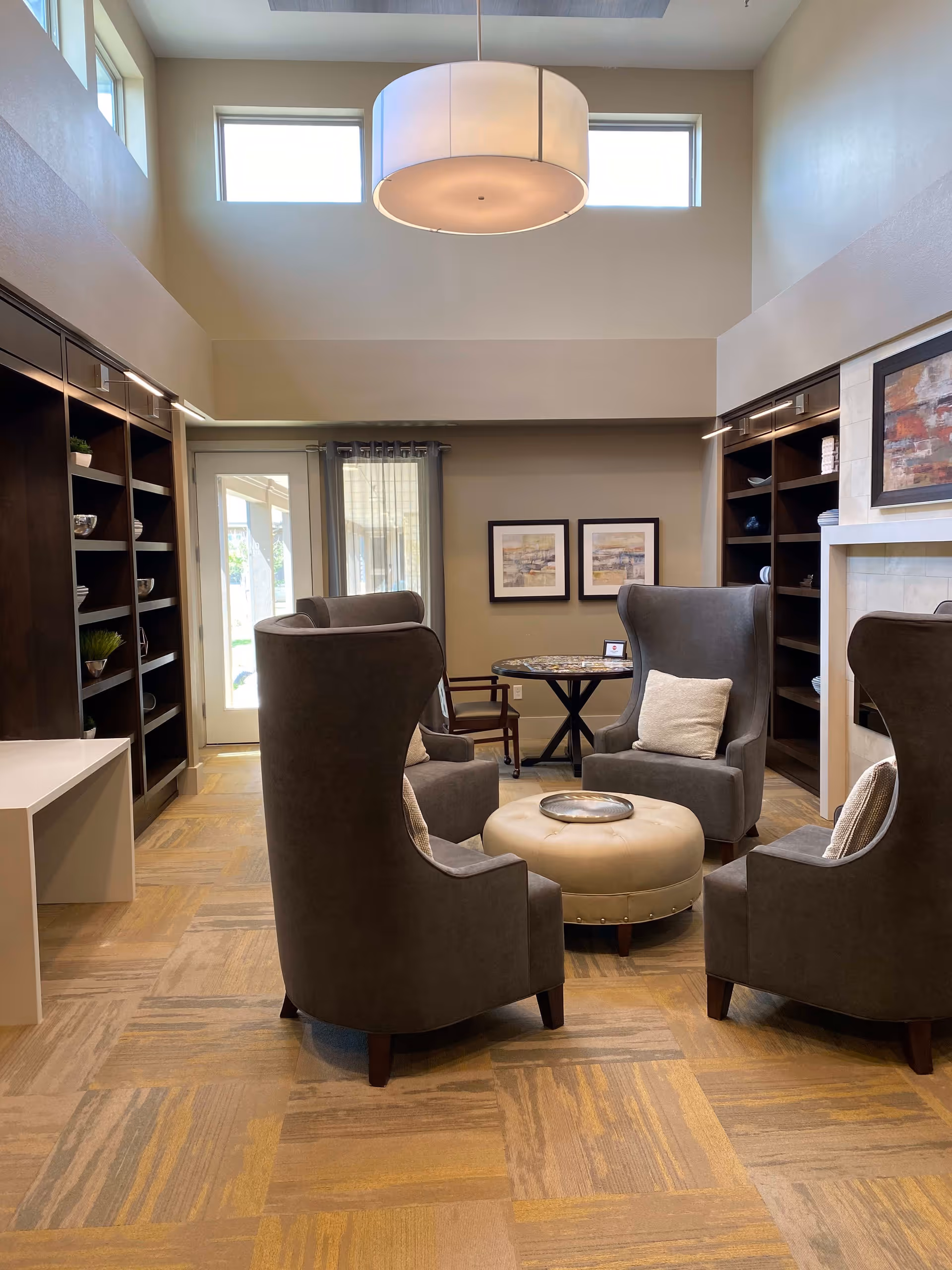 A cozy sitting area with four high-backed gray armchairs arranged around a round beige ottoman with a silver tray on top. The room has a high ceiling with two small windows near the top and a large round hanging light fixture. There are built-in dark wood shelves on both sides of the room, some decorated with small plants and bowls. A fireplace with a framed abstract painting above it is visible on the right side. Two framed pictures hang on the far wall above a small round table and a wooden chair near a glass door with sheer curtains.