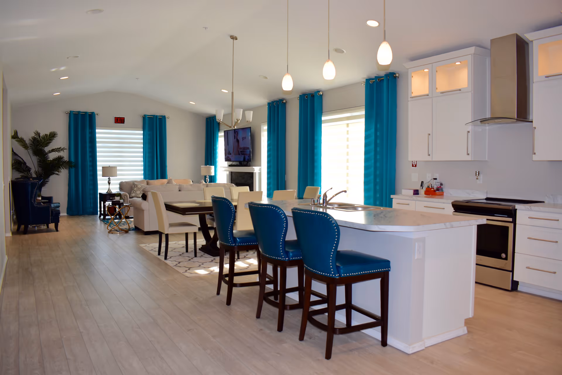 Open-plan bright interior with a kitchen island, turquoise barstools and curtains, and a living area in the background.