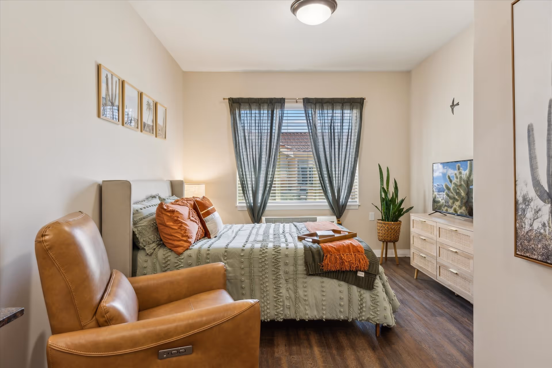 A cozy bedroom with a bed dressed in green bedding and orange pillows, a brown leather armchair, a window with dark curtains, a wooden dresser with a TV on top, a potted plant, and framed pictures on the walls.