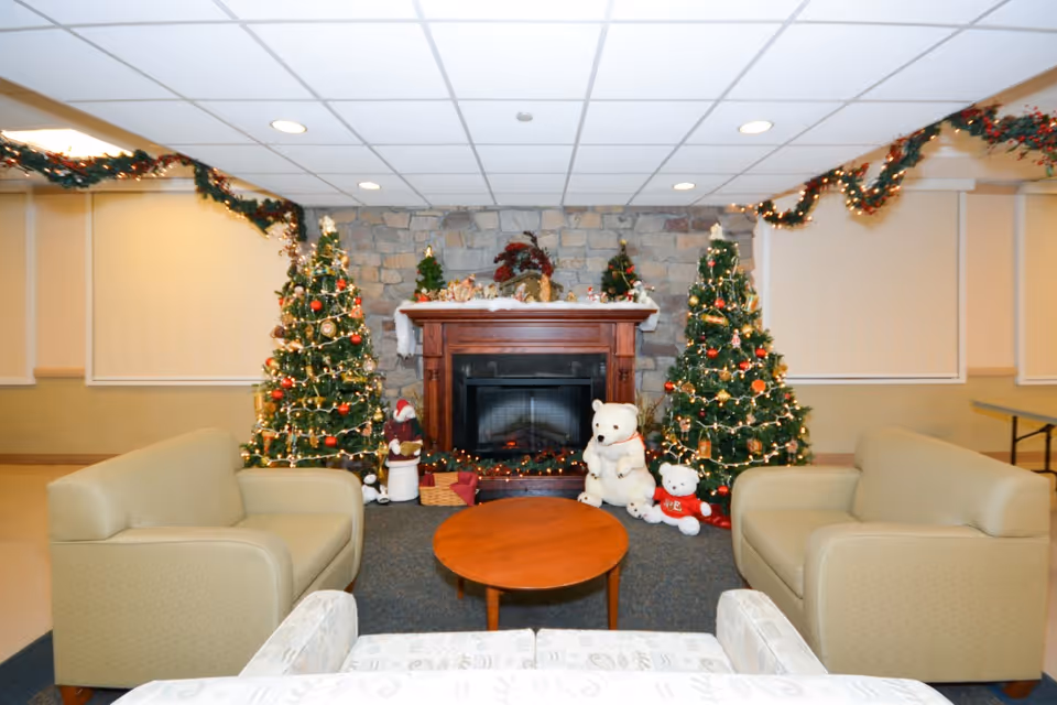 A cozy common area decorated for Christmas with two decorated Christmas trees on either side of a stone fireplace. There are two beige armchairs facing a round wooden coffee table, and a patterned sofa in the foreground. The fireplace mantel is adorned with holiday decorations, and garlands with lights hang along the ceiling edges.